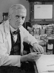 Photograph of Alexander Fleming in a white laboratory coat, seated at his bench with glassware behind him